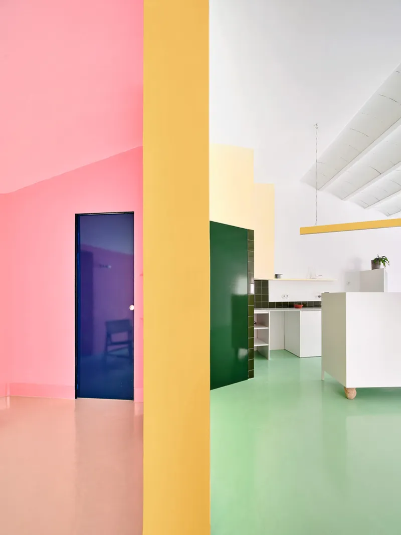 View through yellow column dividing pink floored hallway from green floored kitchen with dark blue door