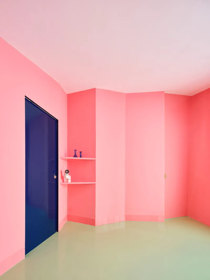 Corner room with pink gradient walls, pale green flooring, corner shelves, and a dark blue door
