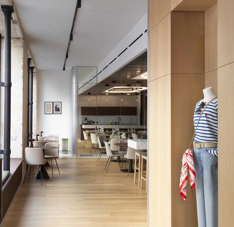 View along blonde wood flooring past exposed stone columns toward cafe seating and kitchen under black track lighting