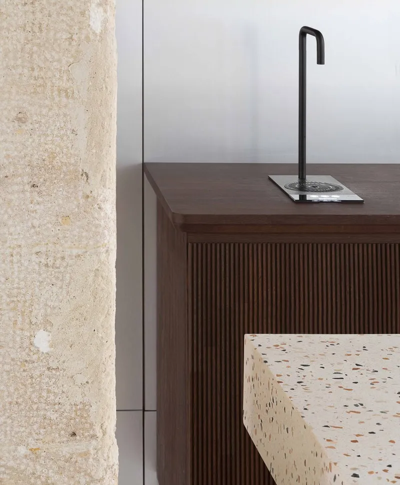 Close-up of matte black faucet on fluted walnut cabinetry beside textured stone column and terrazzo countertop