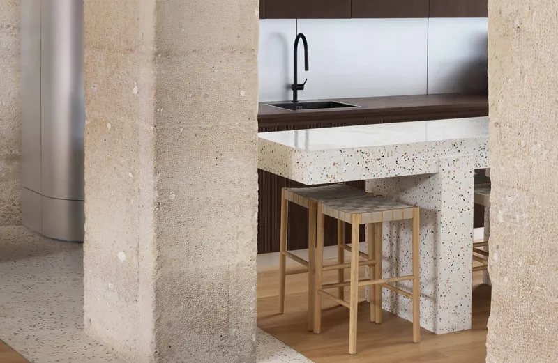 Timber bar stools tucked beneath terrazzo island framed by two rough stone columns
