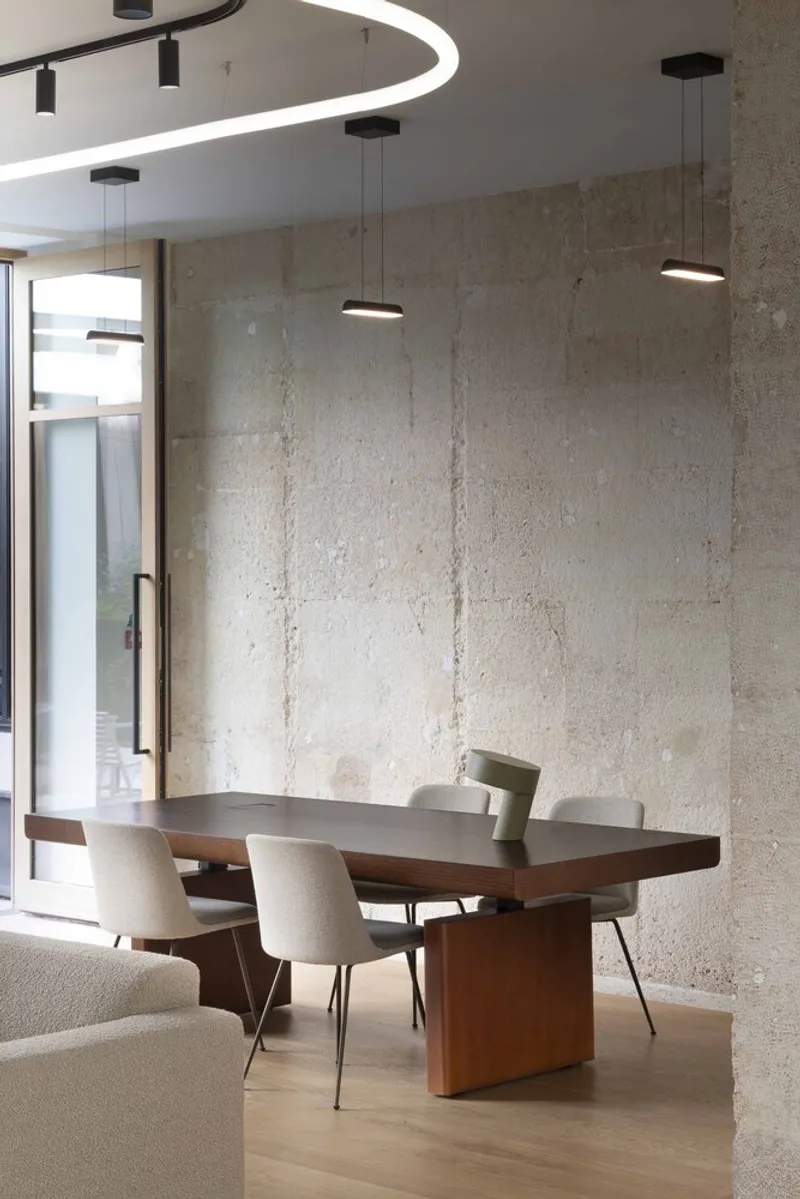 Dining area with walnut table beneath pendant lights and curved overhead fixture against textured concrete wall