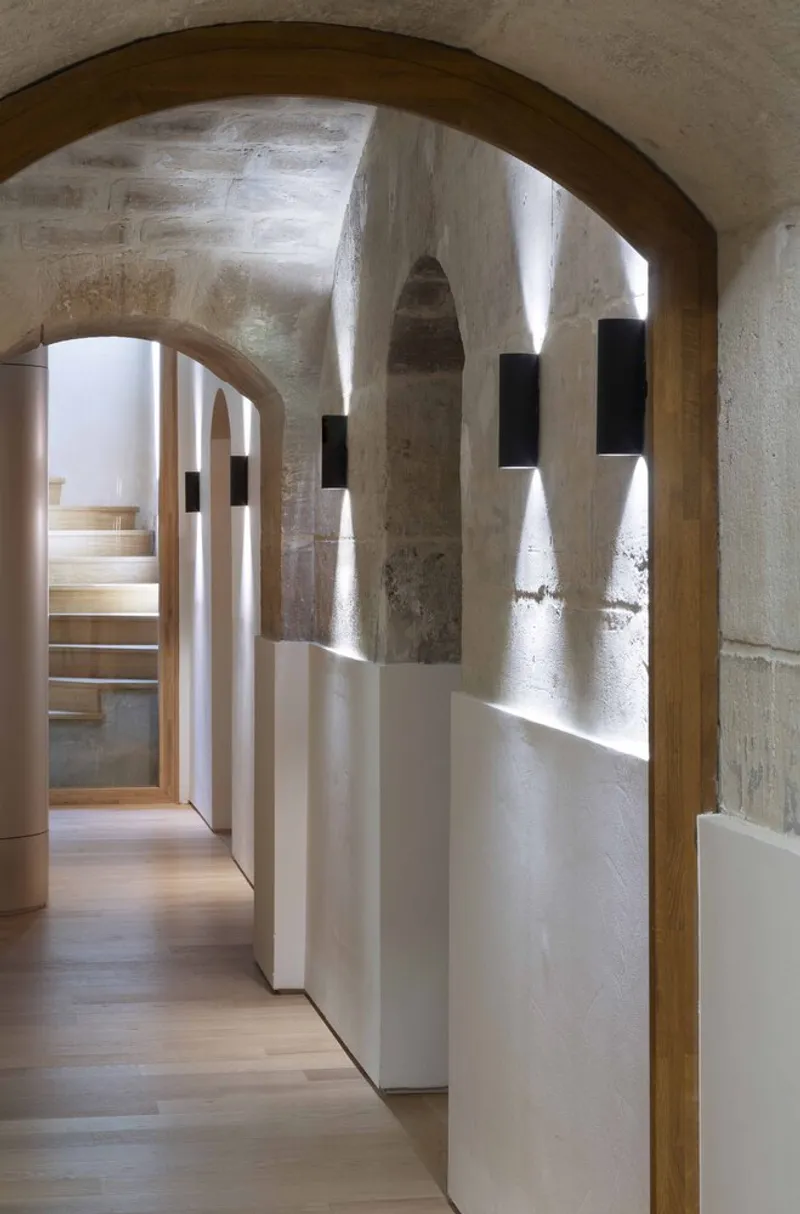 Vaulted passage with arched doorways and wall-mounted sconces illuminating white partitions and concrete columns