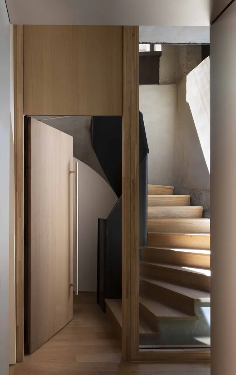 Timber-framed stairwell with illuminated treads rising alongside concrete wall and narrow corridor