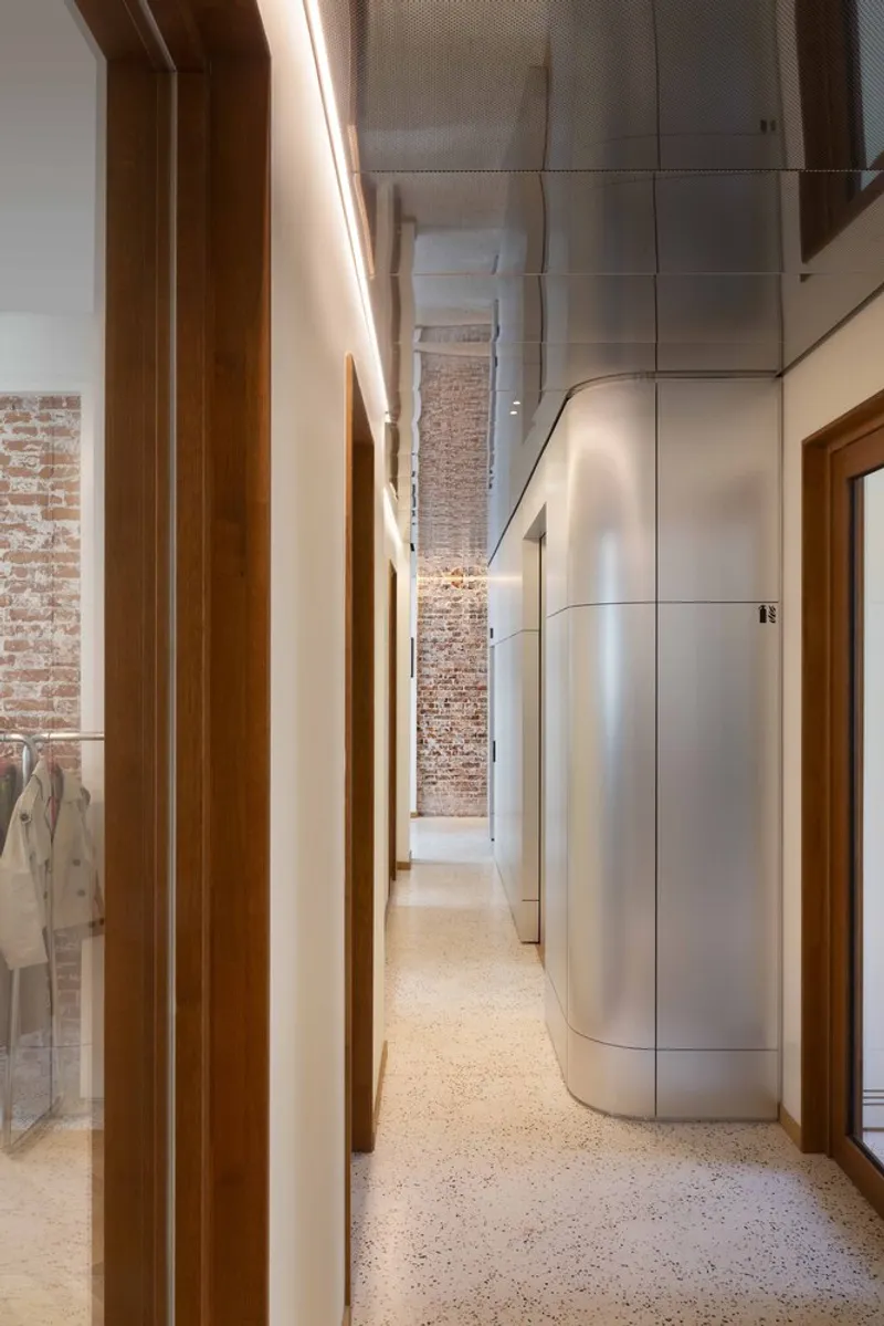Narrow corridor with curved metal panels alongside exposed brick and timber doorways under linear lighting