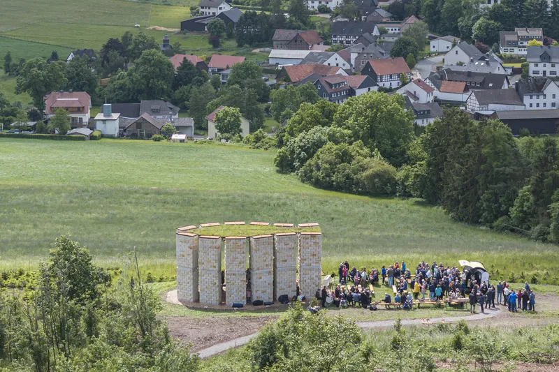 From across the valley: chapel ring of sandstone columns with people gathered for the opening, village behind