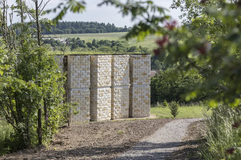 Exterior detail: stack of golden-cream sandstone blocks forming a wall with niche openings, gravel path, trees