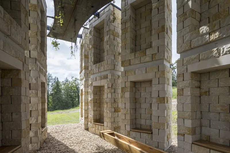 Inside between columns: rough-cut sandstone walls with deep niches, timber bench at the base, view to landscape through the gaps