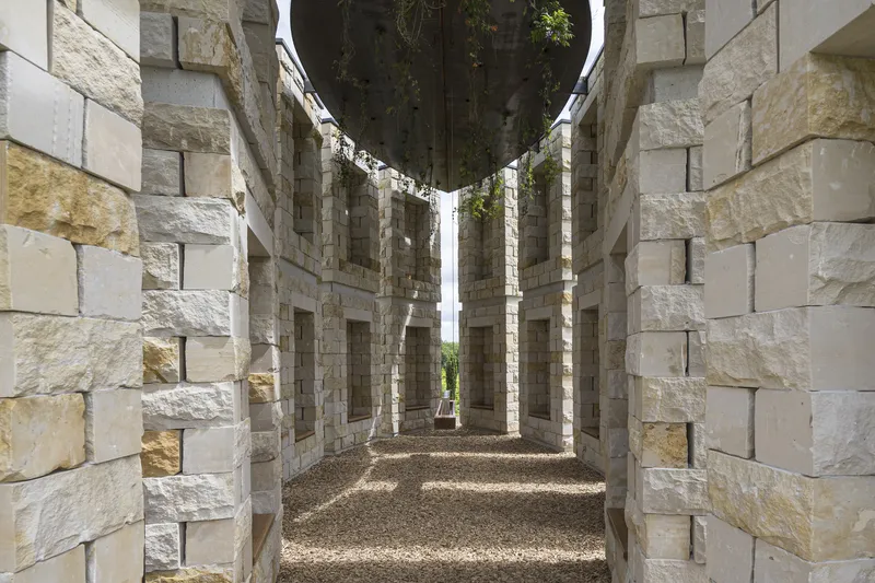 Looking down a corridor between two rows of stone columns with niches, gravel floor, landscape at the end