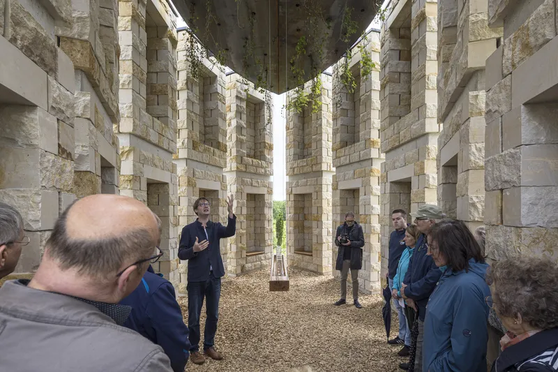 Opening day: group gathered inside the chapel during the inauguration, architect speaking in the centre, stone columns around