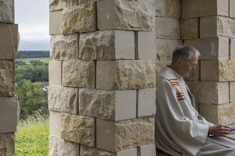 Priest in white robes seated inside reading, sandstone columns behind, view to the landscape