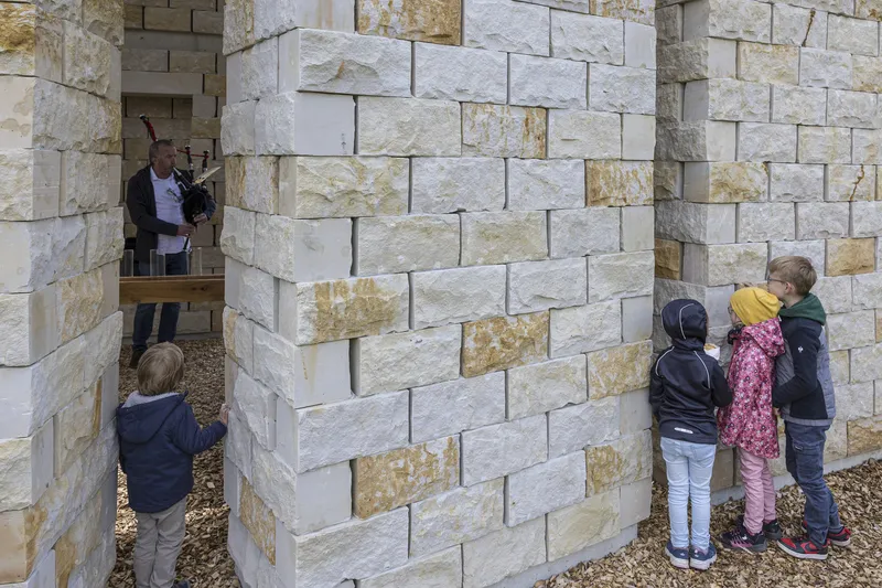 Children peering into a chapel niche, adults visible in another section, gravel ground, sandstone walls