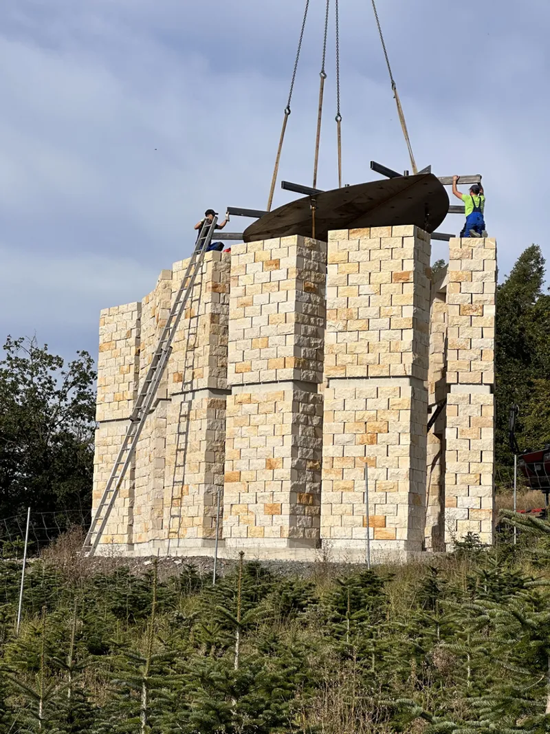 Construction: crane lifting a steel beam over the partially built stone columns, workers on top