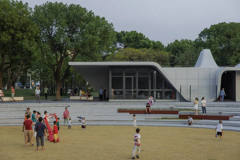 Glazed pavilion entrance with curved roof and terraced plaza where children and families gather on a cloudy day