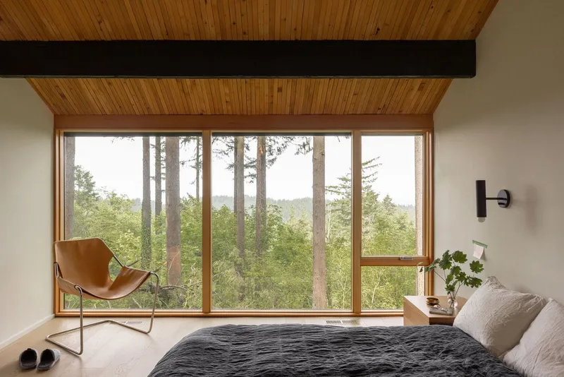 Bedroom with floor-to-ceiling windows framing tall evergreen trees and distant hills