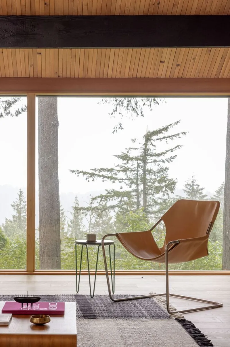 Interior view with leather sling chair and floor-to-ceiling glazing overlooking evergreen trees in mist