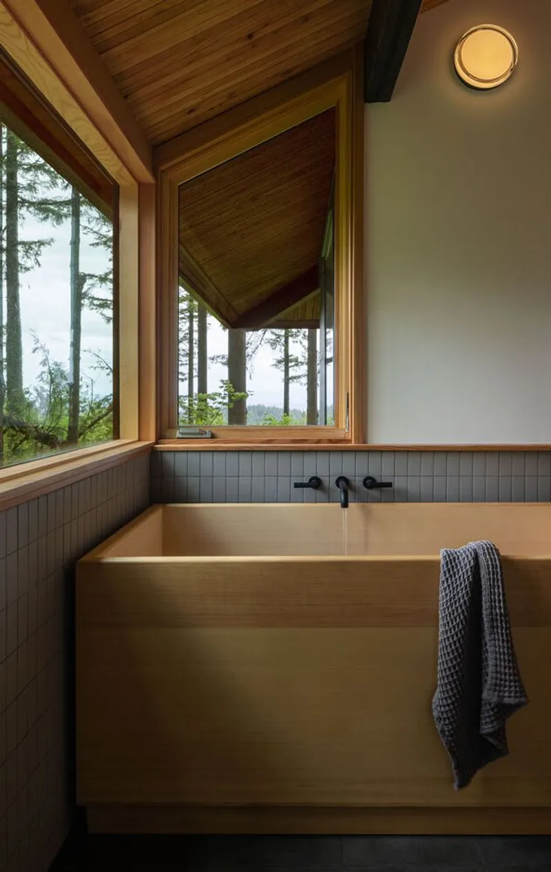Hinoki wood soaking tub beneath corner windows overlooking forested hillside with grey tile wainscoting