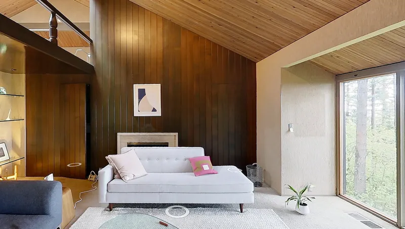 Living area with sloped wood ceiling, dark timber accent wall, white sofa, and forest view through glazing