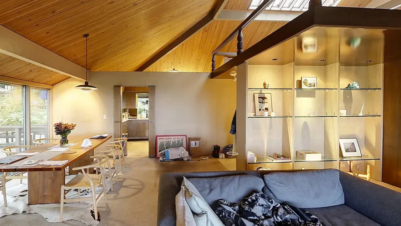 Open-plan living space with exposed timber rafters, backlit shelving unit, and dining table near window