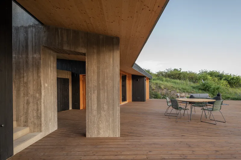 Covered terrace: board-formed concrete columns, CLT ceiling soffit, charred timber walls, round dining table, green meadow view
