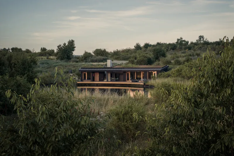 From the meadow: low dark house with timber deck, wildflower planting in foreground, trees, flat Hungarian plain, evening sky