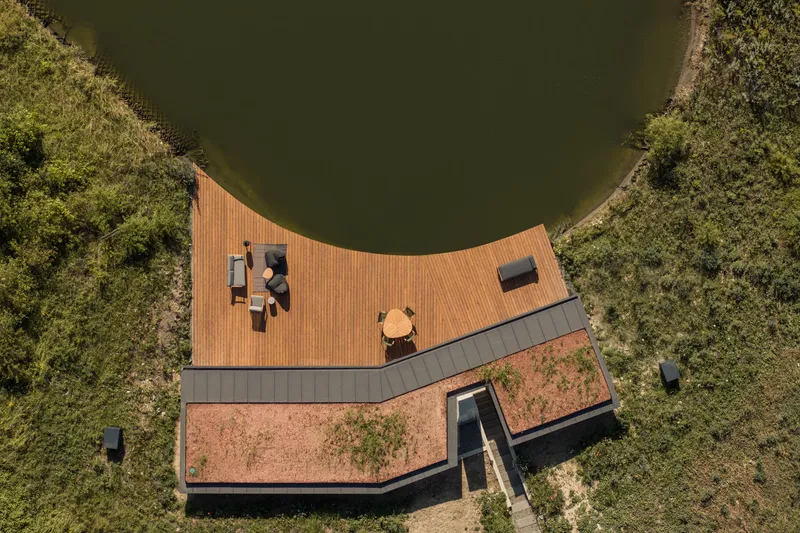 Aerial: V-shaped plan with timber deck wrapping the pond edge, green roof, concrete chimney, meadow surrounding