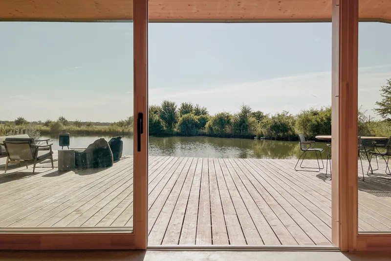 View from inside: timber-framed sliding glass doors, deck, natural pond, chairs, meadow and trees on the horizon