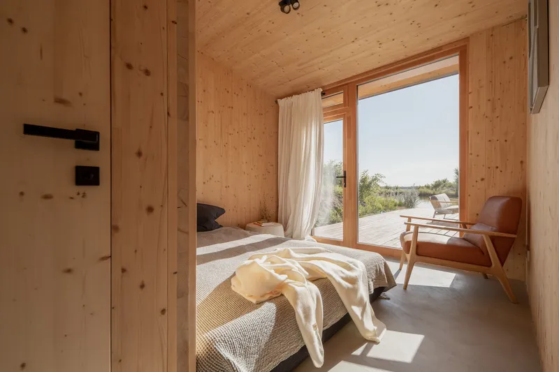 Bedroom: CLT walls and ceiling, timber-framed glass door to the deck, white bedding, leather armchair, sheer curtain