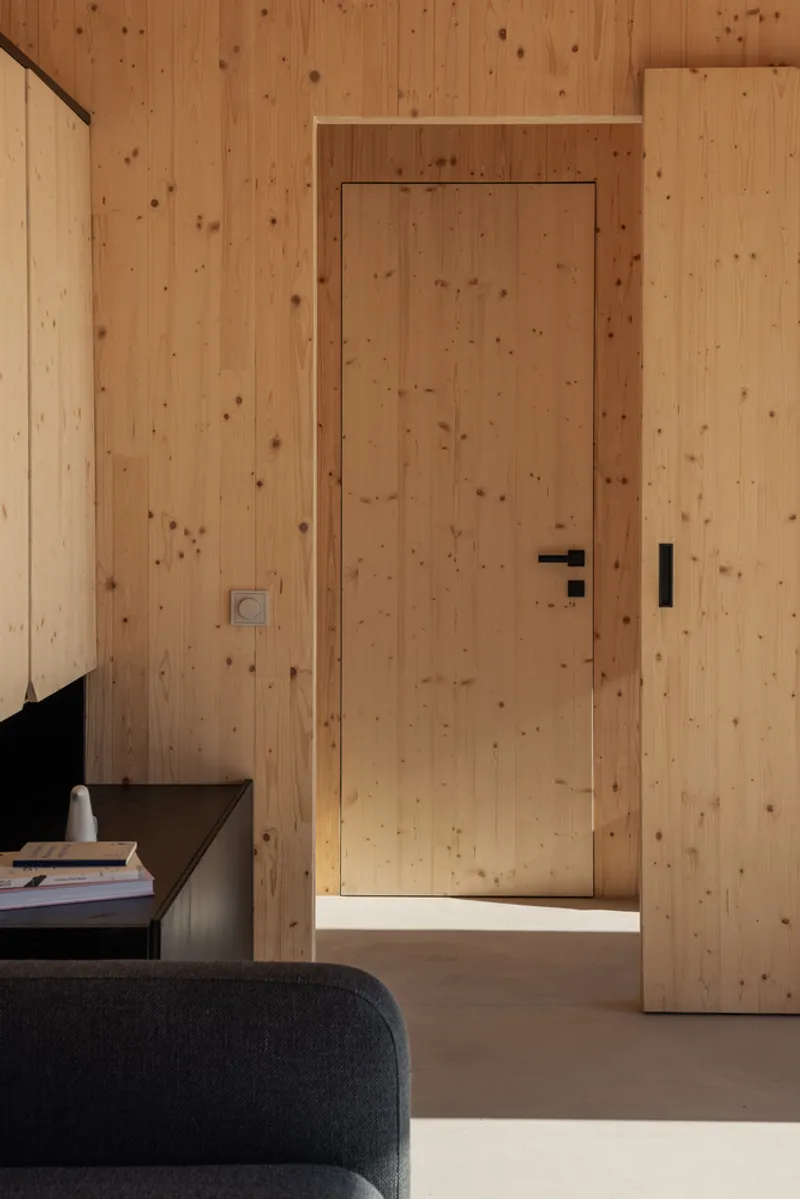 Interior detail: CLT wall with flush timber door, dark sofa edge, afternoon sunlight across the grain