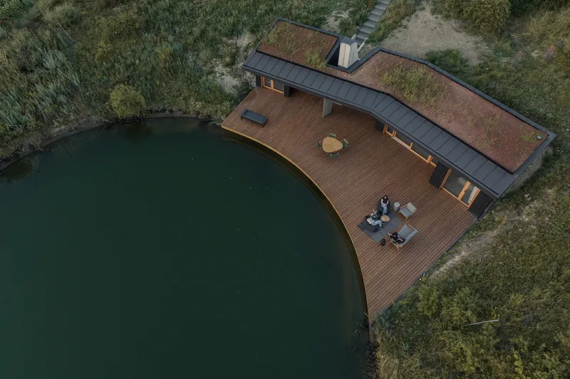 Aerial: V-shaped house with timber deck curving along the pond edge, green roof, two figures sitting on the deck, meadow