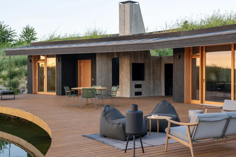Deck and pond: charred timber facade, green roof with grass, concrete chimney, bean bag chairs, fire pit, outdoor furniture, sunset