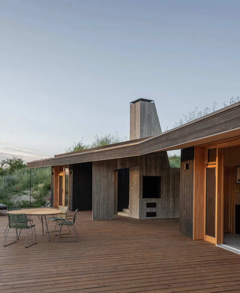 Deck: charred timber wall, concrete chimney with built-in fireplace niche, green metal chairs, green roof above
