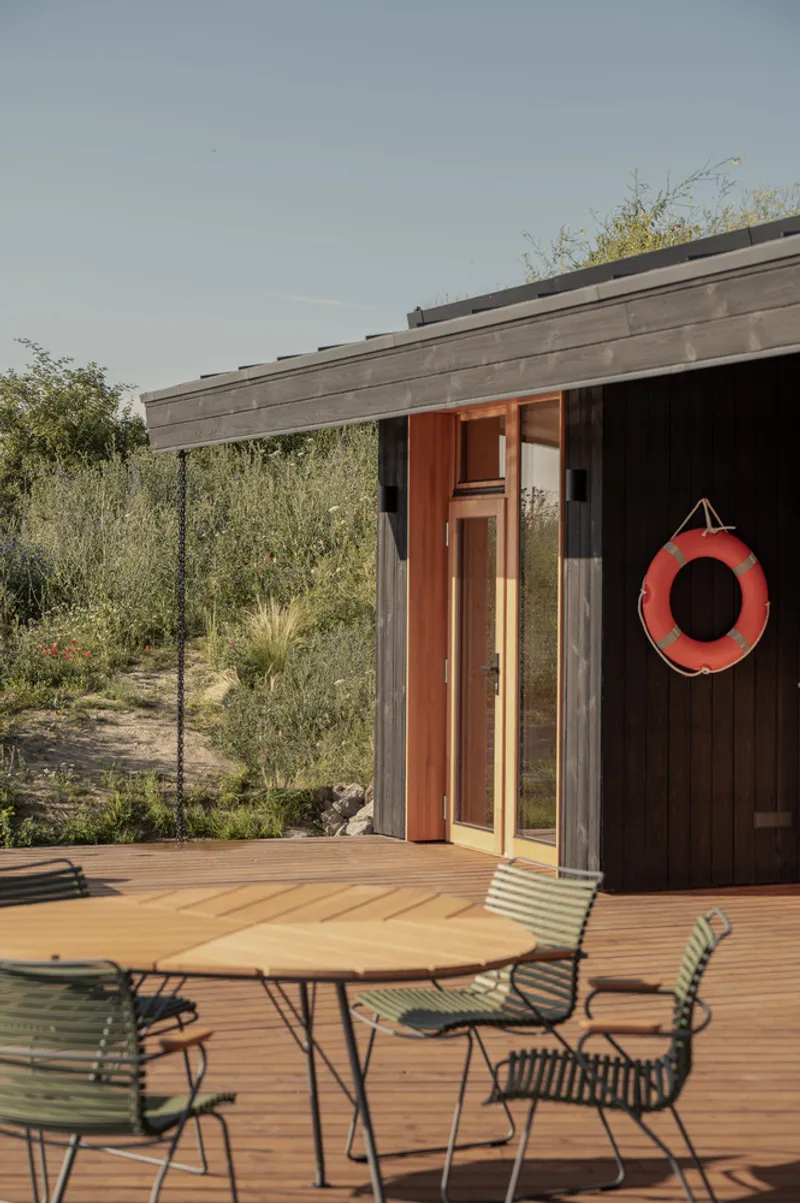 Deck corner: green metal chairs and round table, charred timber wall, orange life ring on hook, meadow grasses