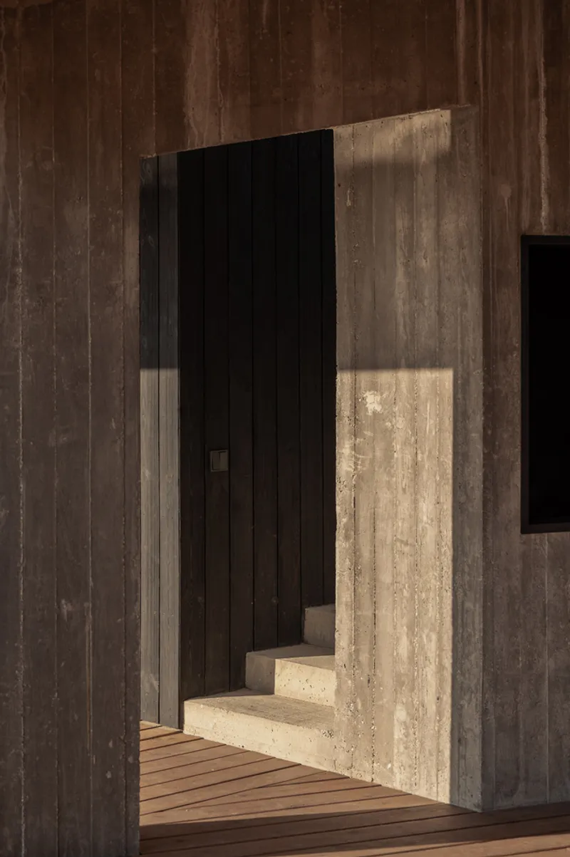 Entry passage: charred timber walls on both sides, concrete step, sharp shadows, timber deck ahead
