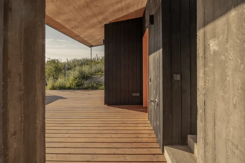Between volumes: CLT ceiling, charred timber cladding, concrete wall, timber deck, meadow visible through the gap
