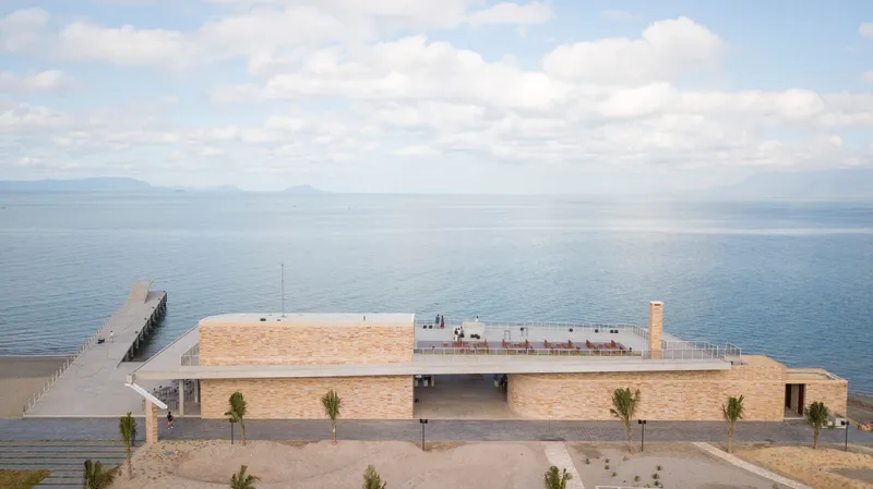 Aerial view of the pale brick waterfront pavilion with a rooftop terrace and timber pier extending into calm waters