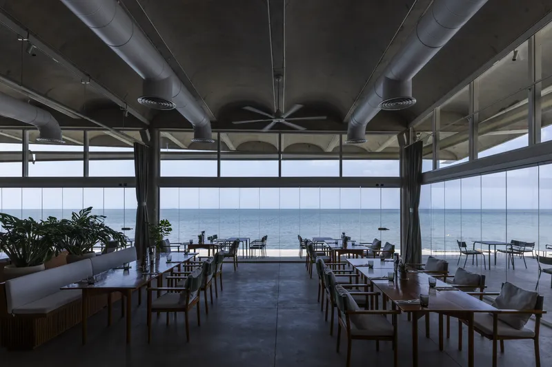 Interior dining space with exposed metal ductwork and floor-to-ceiling windows overlooking the sea