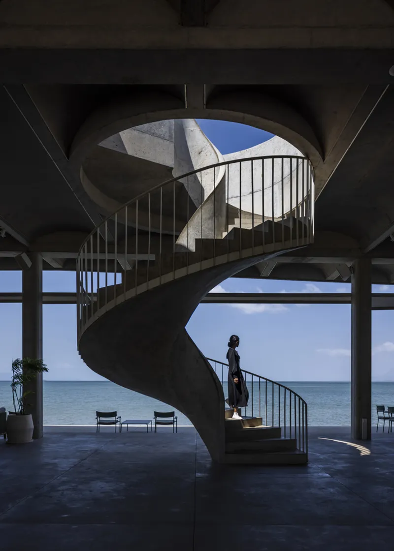 Concrete helical staircase with metal railings framed by vaulted ceiling and ocean view as a figure descends