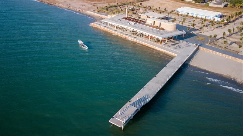 Aerial view of the slatted timber pier extending from the beachfront pavilion into turquoise water with a boat nearby