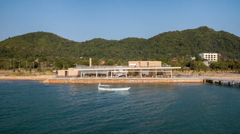 Waterfront view of the low-slung pavilion with exposed concrete columns and brick retaining wall under forested hills