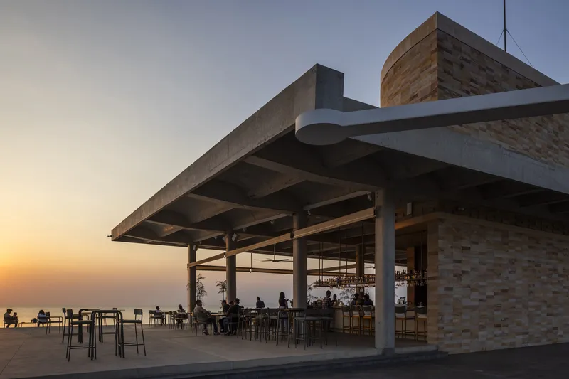 Open-air terrace with concrete canopy and diners seated at tables overlooking the ocean at dusk
