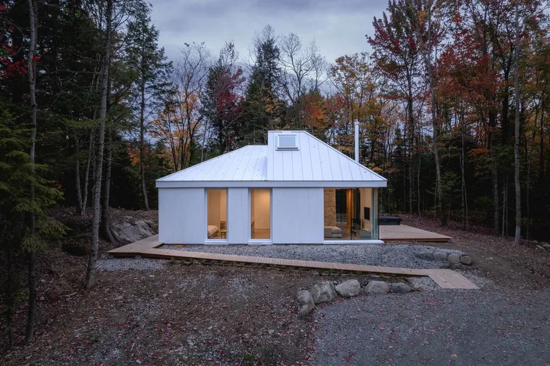 Front facade at dusk: white cube with standing-seam metal roof, timber deck, gravel clearing, autumn forest behind
