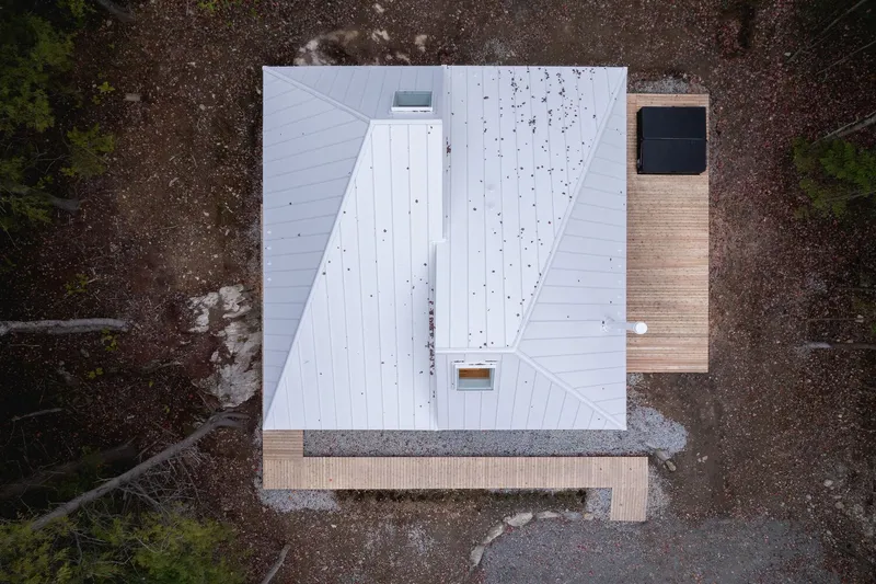 Aerial plan view: white standing-seam roof with twin inverted slopes, square skylights, timber deck, forest clearing