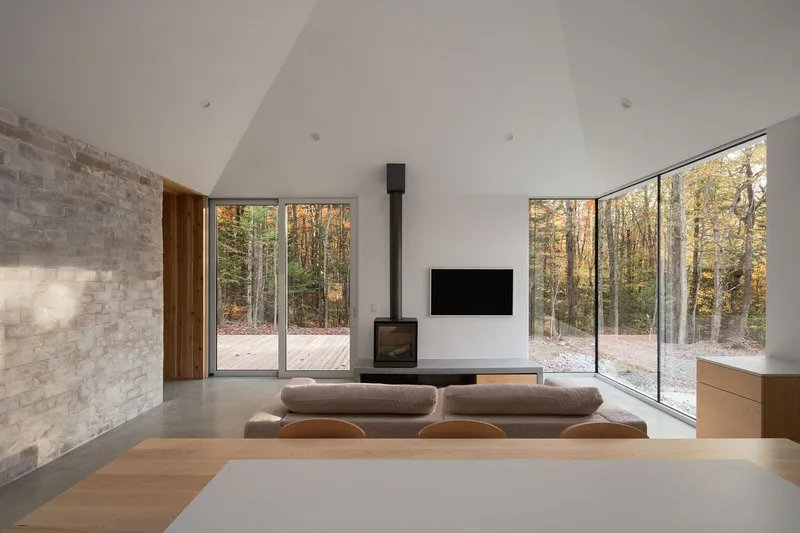 Living room: natural stone feature wall, black wood stove, floor cushions, polished concrete floor, full-height glass to forest