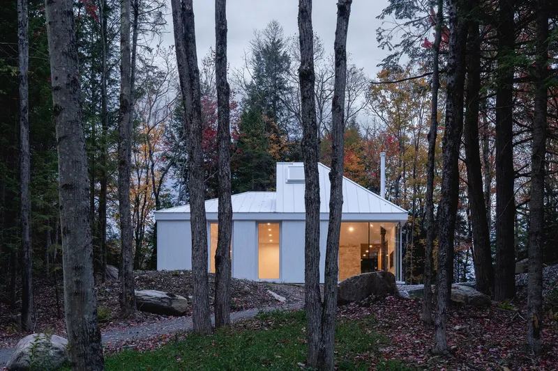 Through the trees at dusk: white house with stone wall visible through glass, boulders, warm interior light