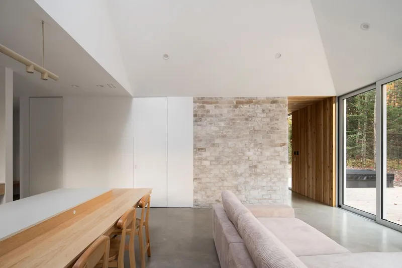 Living and dining: stone wall, oak dining table with chairs, sofa, full-height glass on two sides, forest view