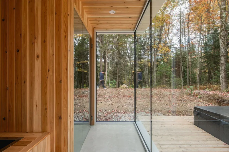 Sauna entry: cedar-clad walls and ceiling, floor-to-ceiling glass corner, timber deck, forest view beyond