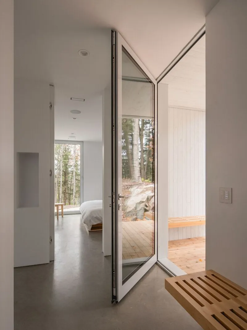 Pivot door: open between corridor and bedroom, white walls, timber slatted bench, forest light