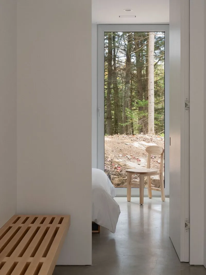 Bedroom through doorway: floor-to-ceiling glass, wooden stool, white bedding, conifer trunks outside
