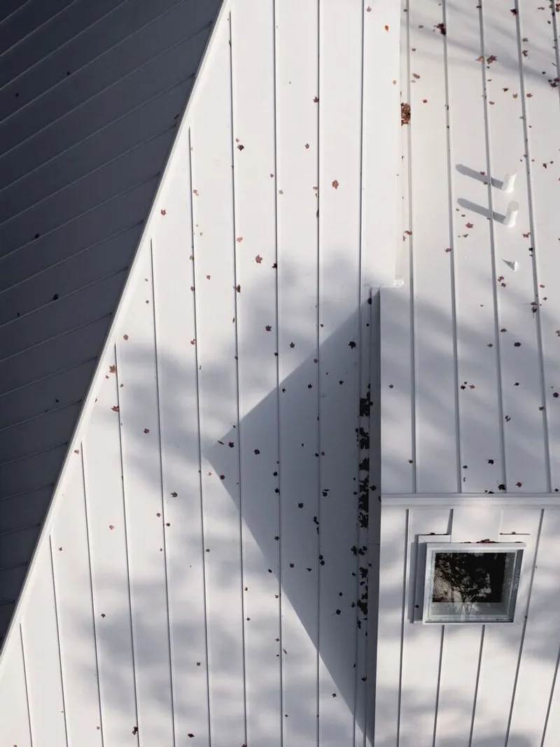 Aerial roof detail: white standing-seam metal, square skylight casting shadow, fallen autumn leaves on panels